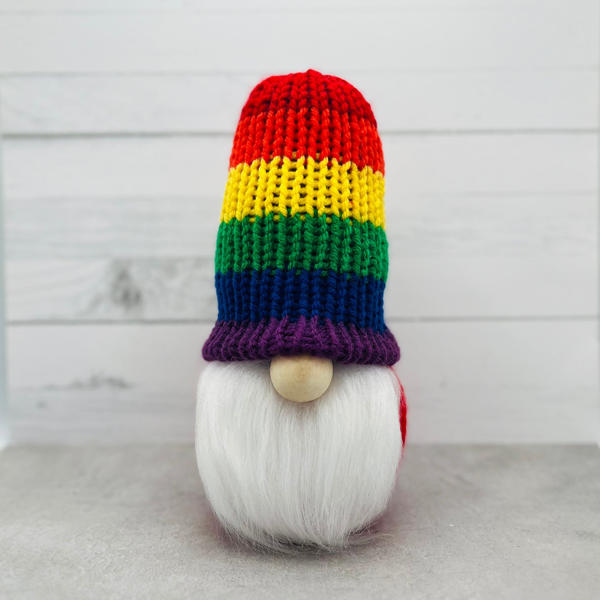 An 8" tall gnome with a rainbow hat with horizontal stripes starting with red at the top then orange, yellow, green, blue and finally purple.  He has a 1" round wood bead for his nose and a white faux fur beard.  His body and hat are knit with yard and filled with dried beans and poly stuffing.