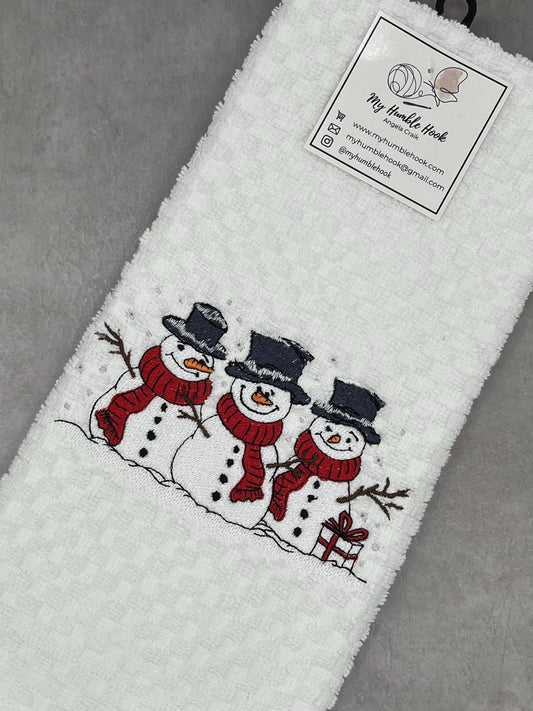 A white kitchen towel embroidered with 3 snowmen.  Each snowman is wearing a red scarf and a dark gray top hat